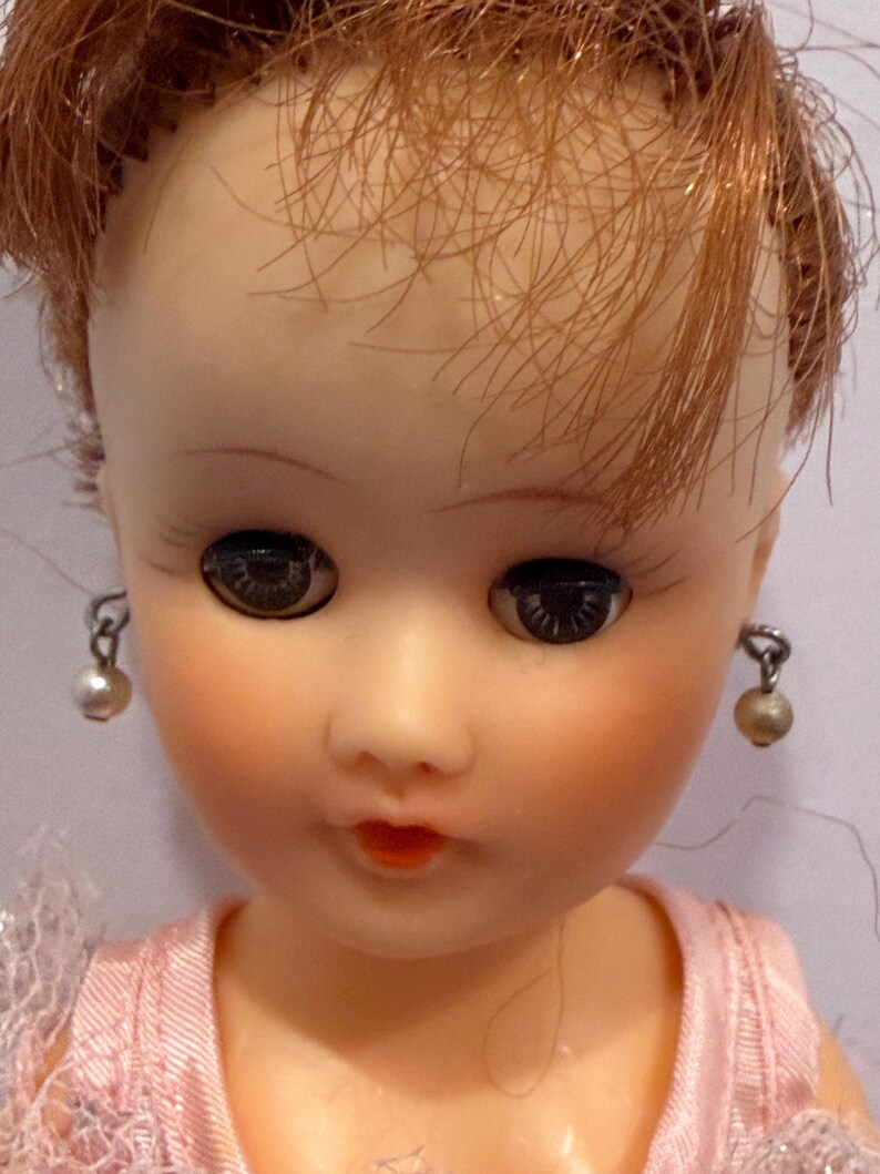 May include: Close-up of a vintage doll's face. The doll has red hair, dark eyes, and is wearing pearl earrings. The doll's lips are painted red, and it is wearing a pink dress.