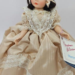 May include: A vintage doll with a cream-colored dress and matching bonnet. The doll has blue eyes, dark hair, and a delicate lace trim. The dress features a full skirt and lace detailing. A tag reads "Madame Alexander Portrait".