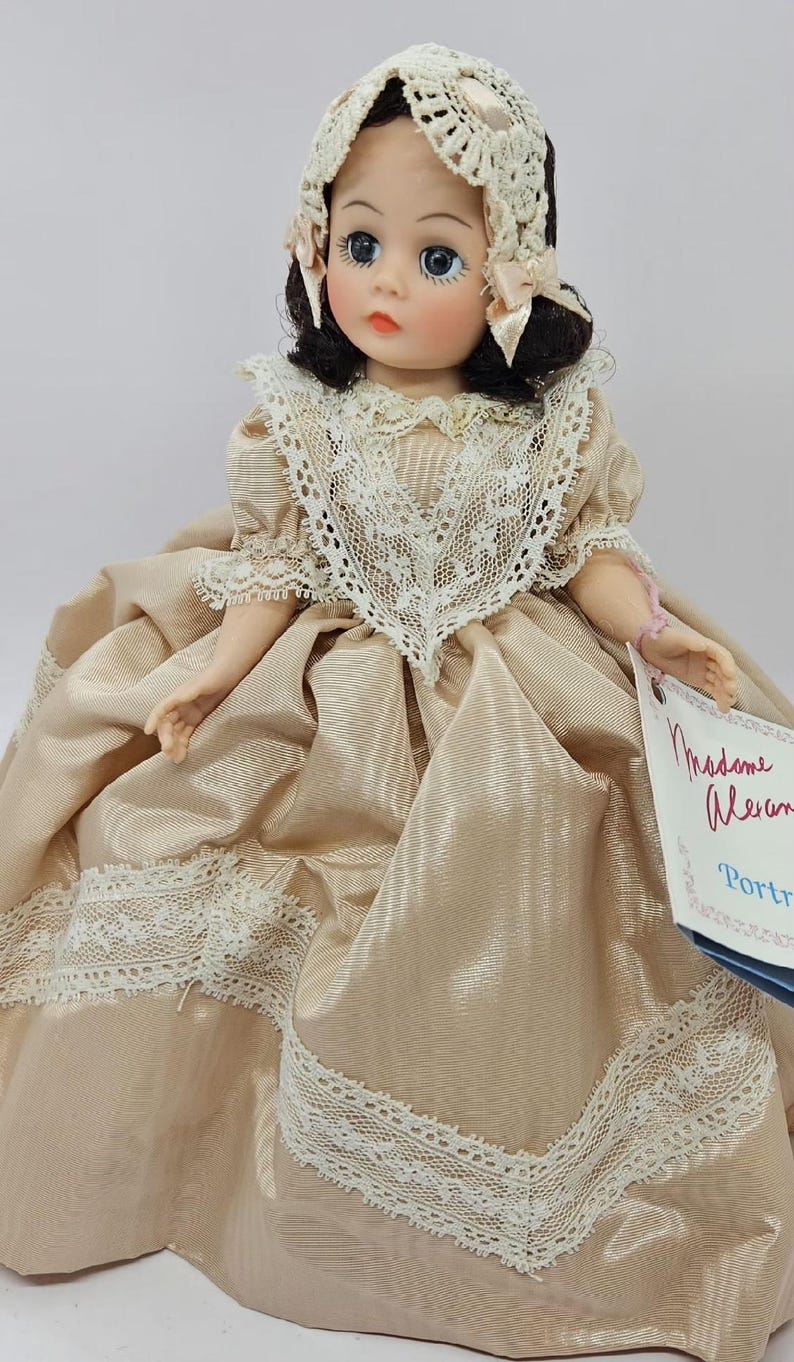 May include: A vintage doll with a cream-colored dress and matching bonnet. The doll has blue eyes, dark hair, and a delicate lace trim. The dress features a full skirt and lace detailing. A tag reads "Madame Alexander Portrait".