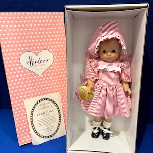 May include: A vintage Effanbee Patsy Joan doll in a box. The doll wears a red and white striped dress, bonnet, and black shoes. The box is pink with a heart pattern and the Effanbee logo. A certificate of authenticity is included.