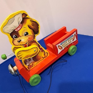 Può includere: Giocattolo vintage in legno da tirare con un cane chef sorridente dei cartoni animati con un cappello bianco e la scritta "TOY TOWN". Il giocattolo è un carretto di hot dog arancione con ruote verdi e la scritta "HOT DOG WAGON".