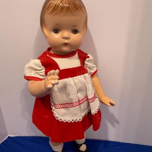 May include: A vintage doll with a painted face, wearing a red dress, white apron, and black shoes. The doll has short, styled brown hair and is posed with one arm slightly raised. The apron has a decorative lace trim.