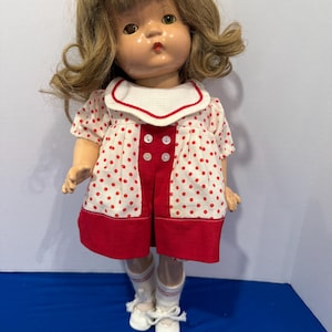 May include: A vintage doll with blonde hair and a red and white polka dot dress. The dress has a white collar and red trim. The doll is wearing white socks and shoes. The doll is a classic toy.