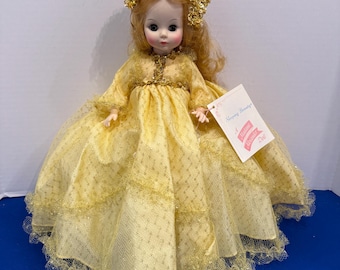 Può includere: Una bambola con lunghi capelli biondi indossa un abito giallo con un copricapo di paillettes dorate. Il vestito presenta una gonna a strati con un sottile motivo a rombi e bordi in pizzo. Un'etichetta bianca riporta Sleeping Beauty A Madame Alexander Doll.