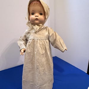Könnte beinhalten: Eine Vintage-Puppe mit geformtem Gesicht, bemalten Gesichtszügen und rotbraunem Haar. Die Puppe trägt ein langärmeliges, beiges Kleid im Leinenstil mit geraffter Taille und eine passende Haube, die mit einem dünnen Stoffband unter dem Kinn gebunden ist.