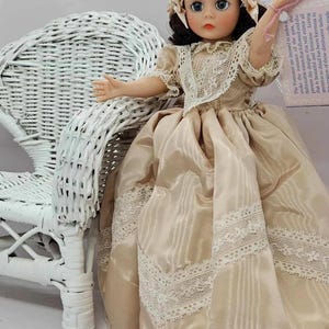 May include: A vintage doll in a cream-colored dress with lace trim and a matching bonnet. The doll has dark hair and blue eyes, and is posed next to a white wicker chair. A tag with text is attached to the doll's wrist.