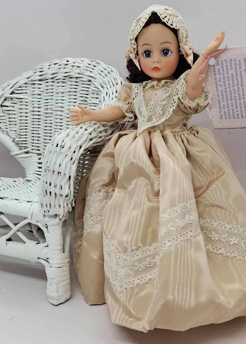 May include: A vintage doll in a cream-colored dress with lace trim and a matching bonnet. The doll has dark hair and blue eyes, and is posed next to a white wicker chair. A tag with text is attached to the doll's wrist.