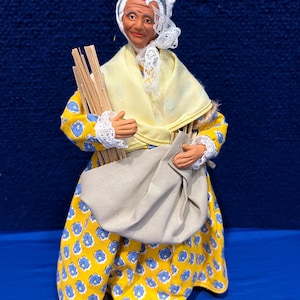 May include: A vintage doll wearing a yellow dress with blue floral print, a white bonnet, and a white lace shawl. The doll is holding a bundle of sticks and a brown sack.