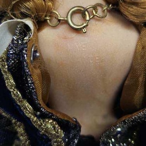 May include: Close-up of a vintage doll's neck and shoulders. The doll has light brown hair styled in curls. A gold-colored chain necklace with a clasp is around the neck. The doll is wearing a dark blue velvet dress with gold trim.