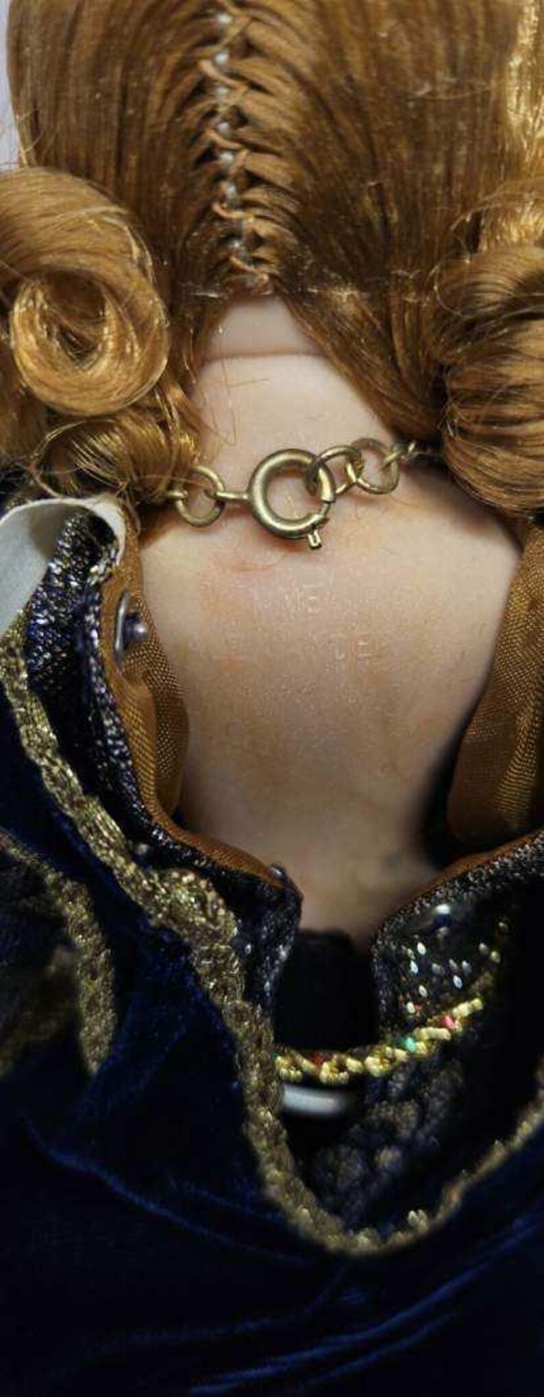 May include: Close-up of a vintage doll's neck and shoulders. The doll has light brown hair styled in curls. A gold-colored chain necklace with a clasp is around the neck. The doll is wearing a dark blue velvet dress with gold trim.