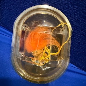 May include: A vintage troll doll with bright orange hair, encased in a clear plastic container with a gold-colored trim. The doll has a small, smiling face and is secured with a yellow string. The background is a deep blue fabric.