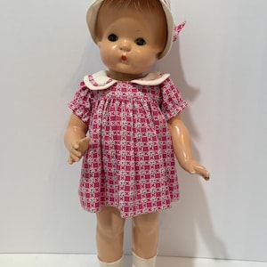 May include: A vintage doll wearing a pink patterned dress with a white collar and matching hat. The doll has white socks and white shoes with gold buckles. The doll's skin is a light peach color.