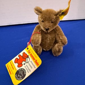May include: A small brown teddy bear with a tag attached. The tag is yellow and has text in multiple languages, including "Für immer Dein" and "Genuine Mohair." The bear has a classic design with a fuzzy texture and small ears.