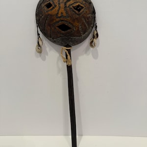 May include: A handcrafted percussion instrument with a round, face-like design. The instrument features a dark wooden handle and a circular head with a carved face design in shades of brown and tan. Small shells hang from the sides.