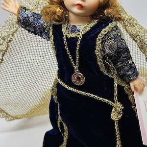 May include: A porcelain doll with green eyes and auburn hair, wearing a dark blue velvet dress with gold trim. The doll has a gold necklace with a red pendant and a gold crown. A gold cape is draped over the doll's shoulders.