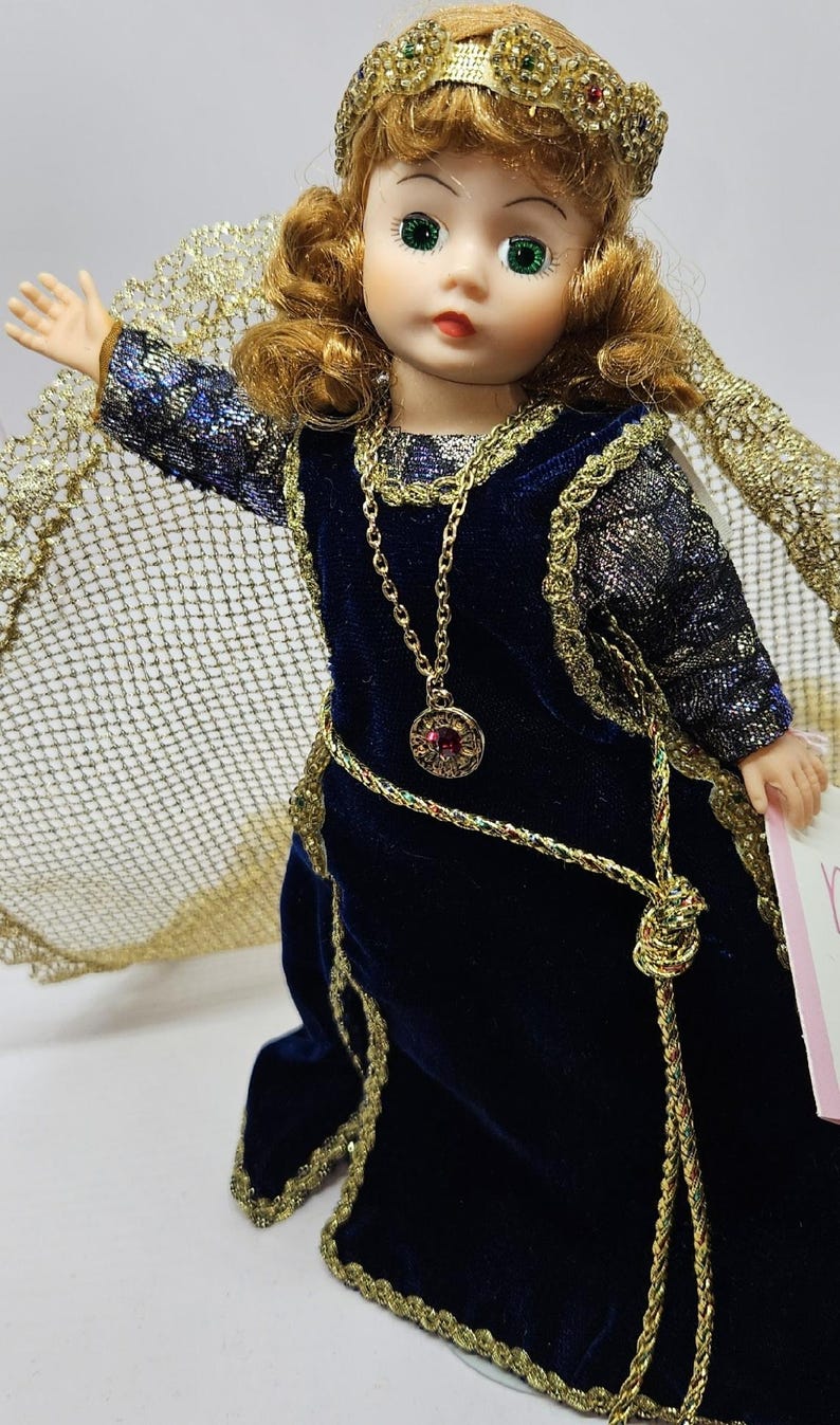 May include: A porcelain doll with green eyes and auburn hair, wearing a dark blue velvet dress with gold trim. The doll has a gold necklace with a red pendant and a gold crown. A gold cape is draped over the doll's shoulders.