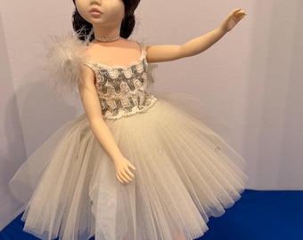 Può includere: Una bambola vintage vestita da ballerina. La bambola ha i capelli scuri e indossa un tutù bianco, un corpetto decorativo e scarpette da ballo rosa. La bambola è in posa con un braccio teso.
