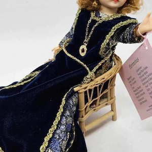 May include: A vintage doll with auburn hair and green eyes, wearing a dark blue velvet dress with gold trim, seated in a small wicker chair. A gold necklace and a tag with text are also visible.