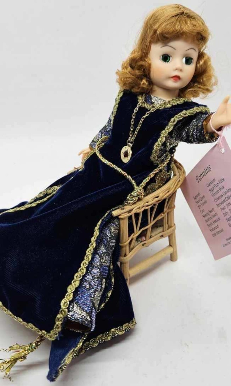 May include: A vintage doll with auburn hair and green eyes, wearing a dark blue velvet dress with gold trim, seated in a small wicker chair. A gold necklace and a tag with text are also visible.