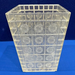 May include: A clear, square vase with a grid pattern and circular floral designs. The transparent material allows light to pass through. The design features squares and circles, creating a geometric and decorative appearance. A decorative item for the home.