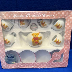 May include: A boxed Kinder-Porzellan-Service tea set. The set includes a teapot, cups, saucers, and a creamer, all decorated with a teddy bear motif. The box is pink with a floral pattern and the words "Kinder-Porzellan-Service".