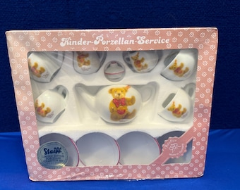 May include: A boxed Kinder-Porzellan-Service tea set. The set includes a teapot, cups, saucers, and a creamer, all decorated with a teddy bear motif. The box is pink with a floral pattern and the words "Kinder-Porzellan-Service".