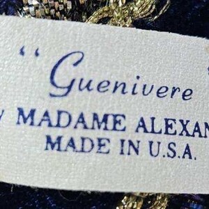 May include: A close-up of a tag with the name "Guenivere" in cursive script. The tag also includes the text "By MADAME ALEXANDER" and "MADE IN U.S.A." in blue ink on a white background. The tag is surrounded by gold and blue fabric.