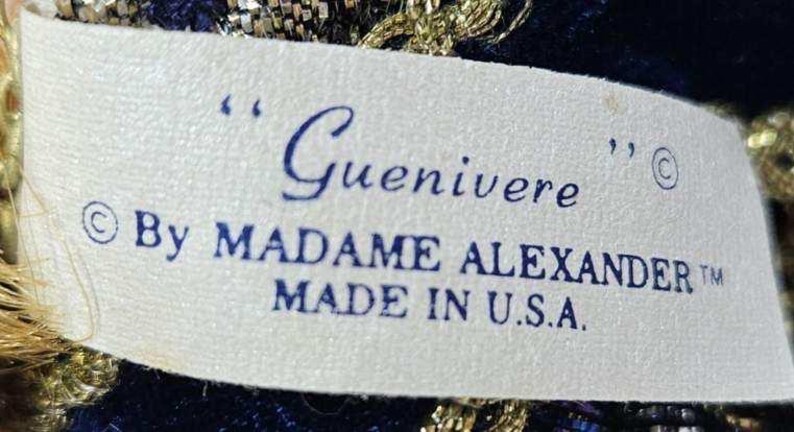May include: A close-up of a tag with the name "Guenivere" in cursive script. The tag also includes the text "By MADAME ALEXANDER" and "MADE IN U.S.A." in blue ink on a white background. The tag is surrounded by gold and blue fabric.