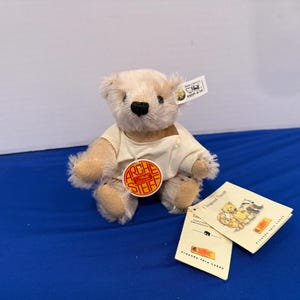 May include: A small, beige teddy bear wearing a white shirt, with a black nose and a round tag that says "ARCHE STEIFF". The bear is sitting on a blue surface, with two small booklets next to it.