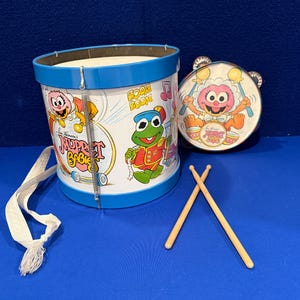 May include: A vintage children's toy drum set featuring characters from The Muppet Show. The set includes a large drum, a tambourine, and two drumsticks. The drum is white with blue trim and has images of Kermit the Frog and other Muppet characters.