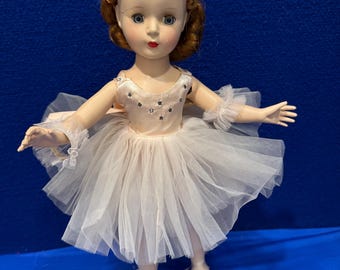 Può includere: Una bambola di porcellana vintage vestita con un costume da ballerina in tulle bianco con un corpetto rosa e dettagli di glitter argentati. La bambola ha i capelli rossi, gli occhi azzurri e le labbra rosse.