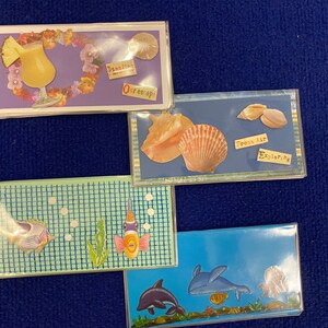May include: A collection of decorative magnets with a tropical theme. The magnets feature images of a pineapple drink, seashells, fish, and dolphins. Some magnets have text that reads "Paradise", "Our escape", "Fresh Air", and "Exploring".