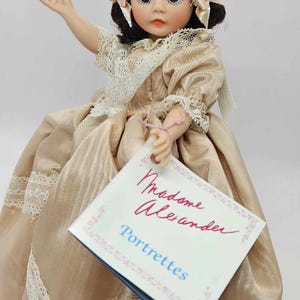 May include: A vintage Madame Alexander Portrettes doll. The doll wears a gold satin dress with lace trim and a matching bonnet. The doll has dark hair, blue eyes, and is holding a tag that reads "Madame Alexander Portrettes".