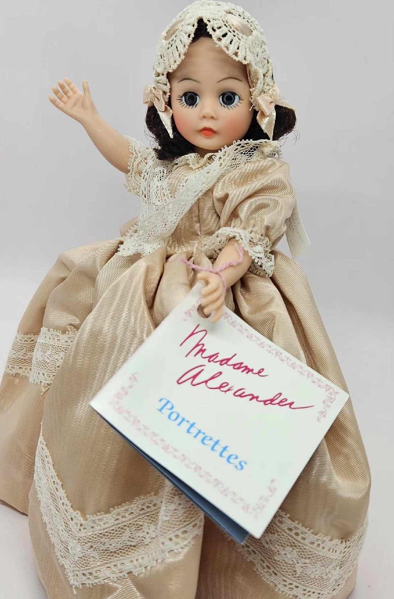 May include: A vintage Madame Alexander Portrettes doll. The doll wears a gold satin dress with lace trim and a matching bonnet. The doll has dark hair, blue eyes, and is holding a tag that reads "Madame Alexander Portrettes".