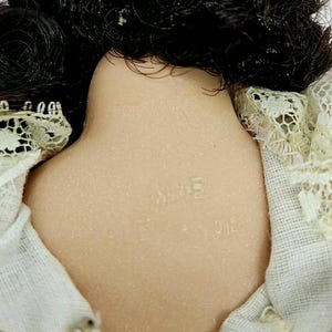 May include: Close-up of a vintage doll's back, showing black hair and a porcelain neck. The doll's neck is stamped with the word "MIME". The doll is wearing a beige dress with lace trim.