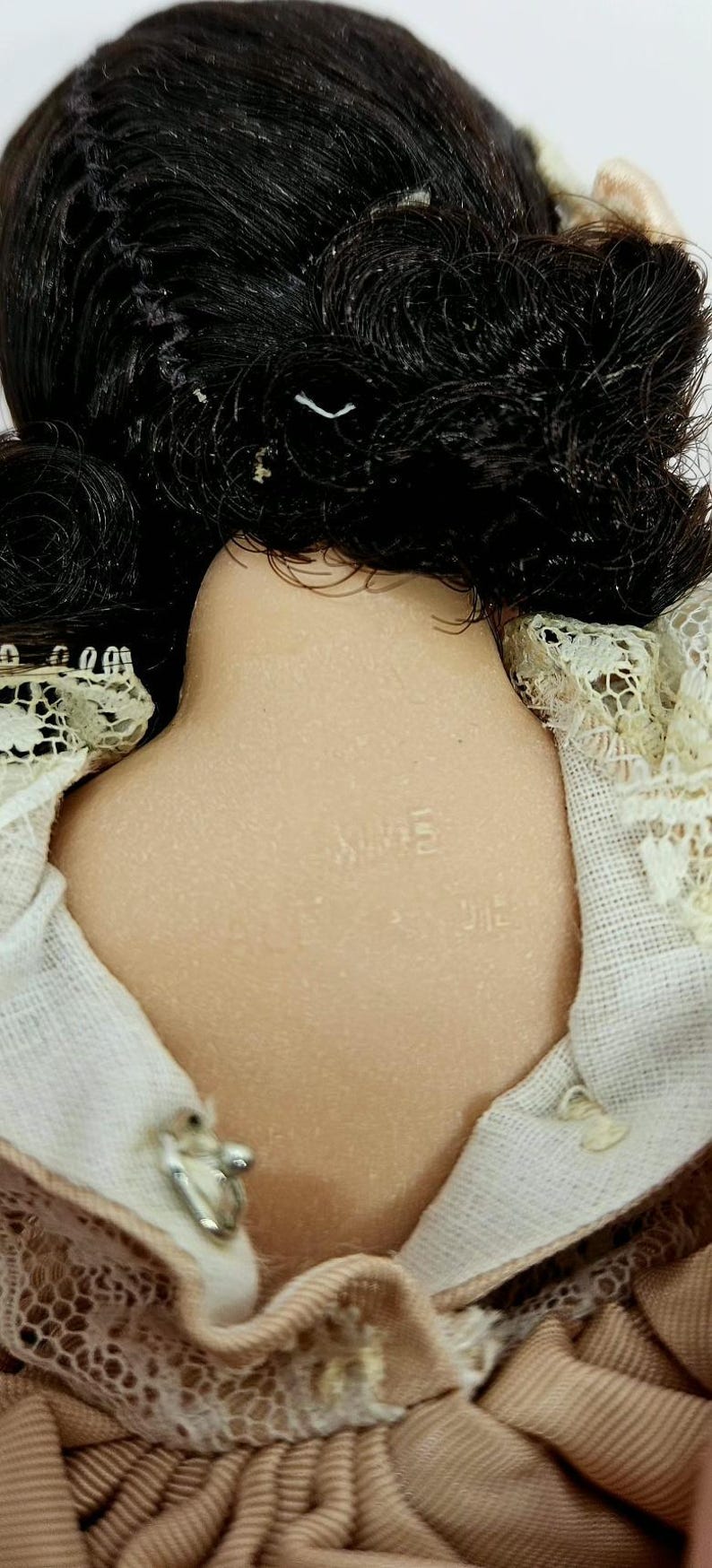 May include: Close-up of a vintage doll's back, showing black hair and a porcelain neck. The doll's neck is stamped with the word "MIME". The doll is wearing a beige dress with lace trim.