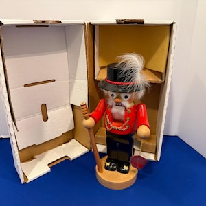 May include: A wooden nutcracker figurine in a red jacket, black pants, and a black top hat with a white feather. The figurine holds a cane and a red rose. It is displayed in an open cardboard box.
