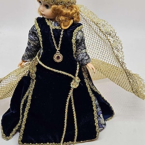 May include: A porcelain doll in a dark blue velvet gown with gold trim and a matching gold headpiece. The doll has a gold necklace with a pendant and a gold net cape. The doll's hair is a light auburn color.