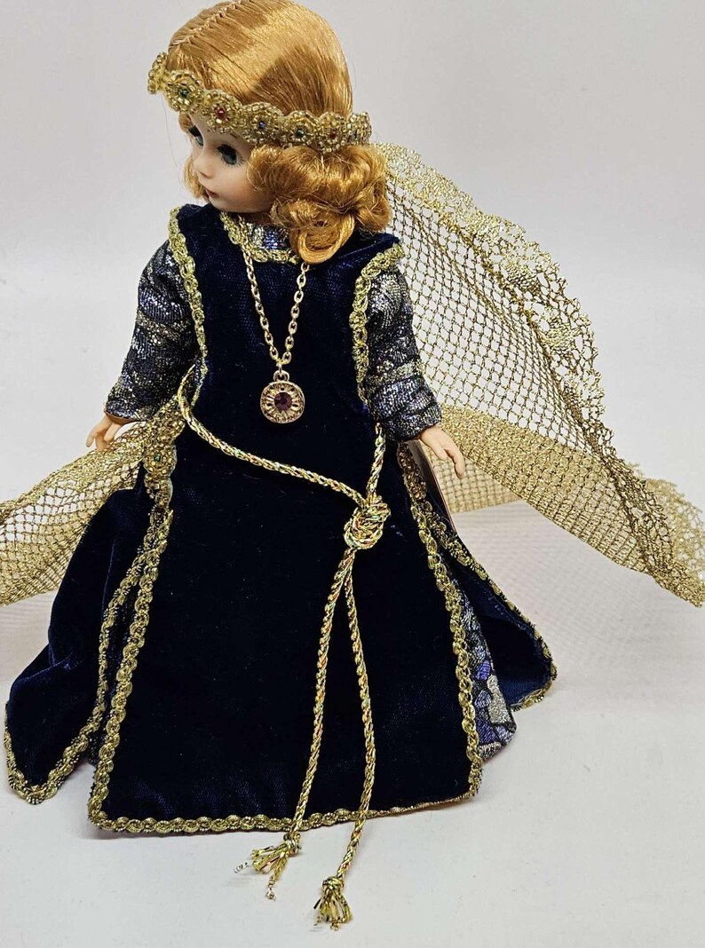 May include: A porcelain doll in a dark blue velvet gown with gold trim and a matching gold headpiece. The doll has a gold necklace with a pendant and a gold net cape. The doll's hair is a light auburn color.