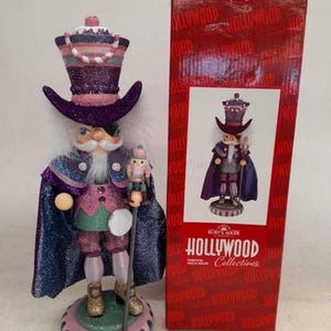 Może przedstawiać: Ozdobna figurka dziadka do orzechów w fioletowo-różowym stroju, z niebieską peleryną i wysokim kapeluszem. Figurka trzyma laskę i miniaturowego dziadka do orzechów. Podstawa jest różowo-zielona. Obok czerwone pudełko z napisem "Hollywood Collections".