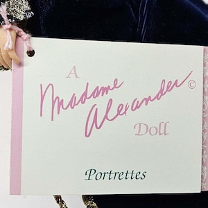 May include: A Madame Alexander doll tag with the words "A Madame Alexander Doll Portrettes" in pink script. The tag is attached to a doll's hand and a dark blue velvet dress.