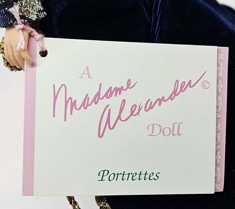 May include: A Madame Alexander doll tag with the words "A Madame Alexander Doll Portrettes" in pink script. The tag is attached to a doll's hand and a dark blue velvet dress.