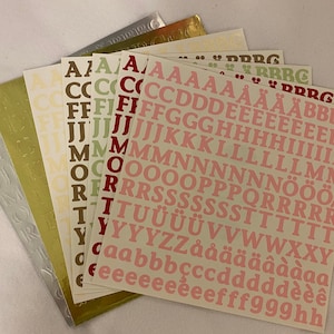 May include: A set of alphabet stickers in various colors, including red, green, brown, and pink. The stickers are arranged in rows, with each row containing a different letter of the alphabet. The stickers are on a white background.
