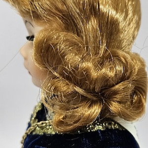 May include: A porcelain doll with golden-brown hair styled in an updo. The doll is wearing a dark blue velvet dress with gold trim. The dress has a detailed, patterned design on the sleeves and bodice. The doll's profile is visible.