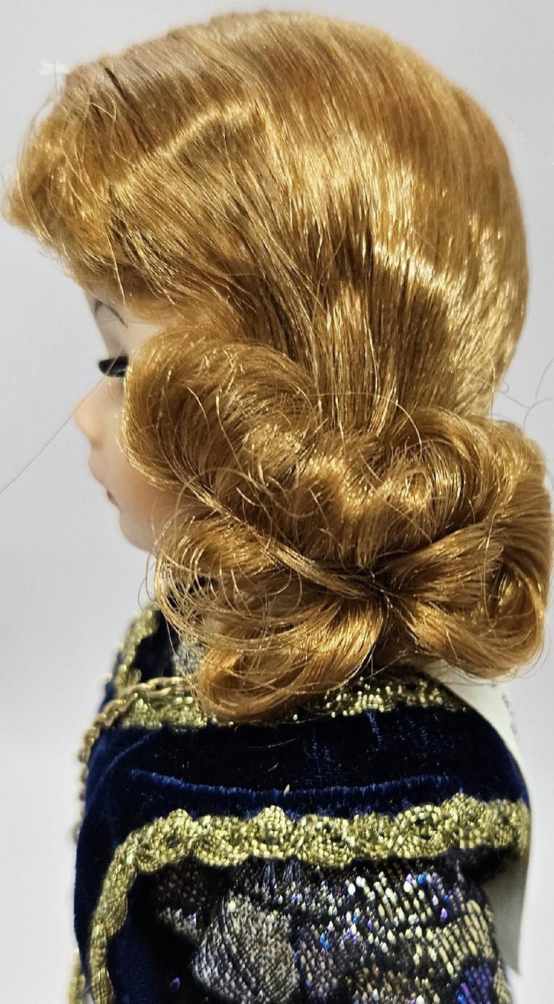 May include: A porcelain doll with golden-brown hair styled in an updo. The doll is wearing a dark blue velvet dress with gold trim. The dress has a detailed, patterned design on the sleeves and bodice. The doll's profile is visible.