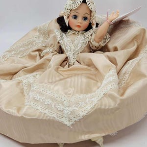 May include: A vintage doll with a porcelain face, wearing a cream-colored dress with lace trim and a matching bonnet. The doll has dark hair, blue eyes, and a delicate pink bracelet. The dress is gathered at the waist and flares out.