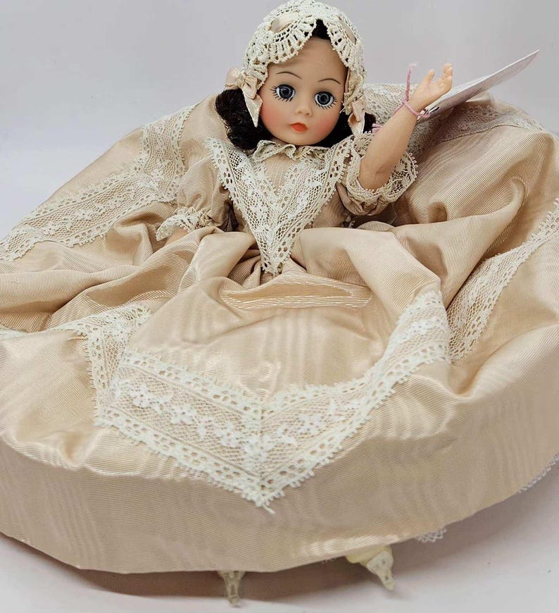 May include: A vintage doll with a porcelain face, wearing a cream-colored dress with lace trim and a matching bonnet. The doll has dark hair, blue eyes, and a delicate pink bracelet. The dress is gathered at the waist and flares out.