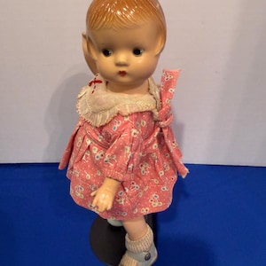 Op de afbeelding: Vintage pop met een geschilderd gezicht, gekleed in een roze bloemenjurk met een witte kanten kraag. De pop heeft roodbruin haar en draagt lichtblauwe schoenen en sokken. De pop staat op een zwarte standaard.