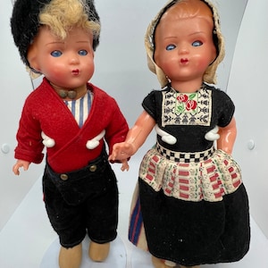 May include: Two vintage dolls dressed in traditional Dutch clothing. The boy doll wears a red jacket, black pants, and a black hat. The girl doll wears a black dress with a white apron and a white bonnet.