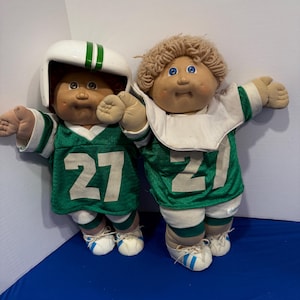 Könnte beinhalten: Zwei Stoffpuppen in grün-weißen Football-Trikots mit der aufgedruckten Nummer 27 auf der Vorderseite. Eine Puppe trägt einen weißen Helm mit grünen Streifen, während die andere lockiges beiges Wollhaar und einen weißen Kragen hat.